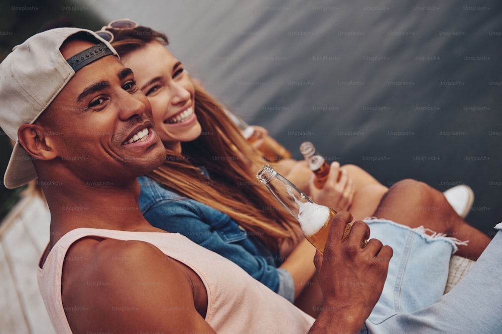 Gruppe glücklicher junger Leute in Freizeitkleidung, die lächeln und Bier trinken, während sie auf dem Pier sitzen