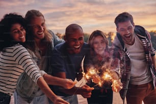 Group of young people in casual wear smiling and holding sparkers while standing on the pier