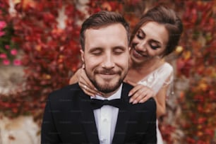 Gorgeous bride gently hugging stylish groom at old wall of autumn red leaves. Happy sensual wedding couple embracing. Romantic moments of newlyweds