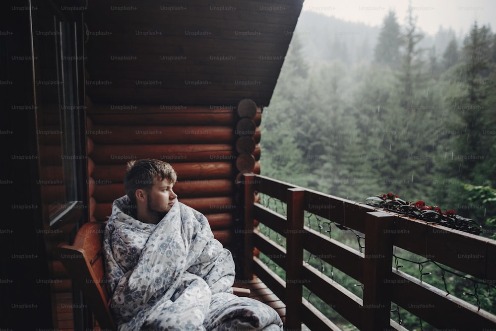 山の森を背景に、雨の日に木造キャビンのポーチでリラックスする毛布のスタイリッシュな男性旅行者。スタイリッシュなヒップスターの休息。テキスト用のスペース。大気モーメント