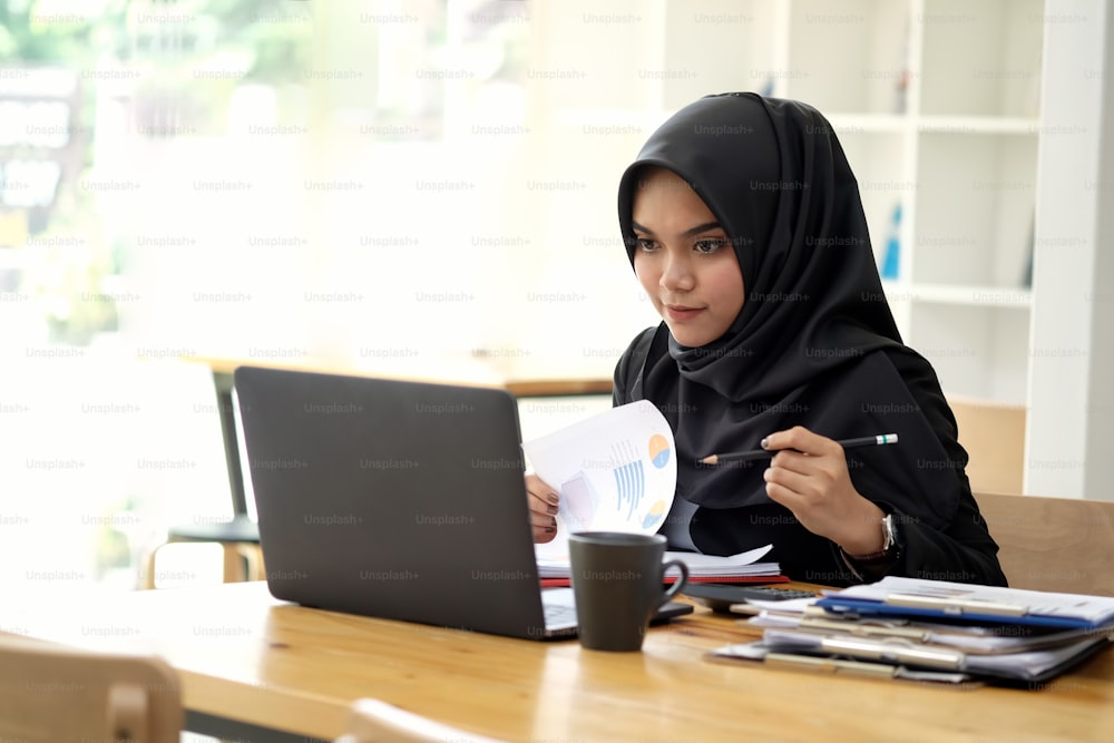 Séduisante jeune asiatique portant un hijab sombre travaillant de la paperasse avec son ordinateur portable.