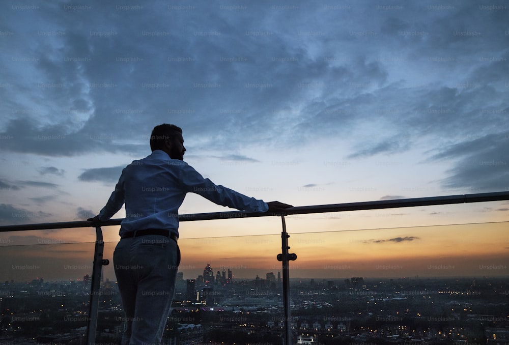 Una vista trasera de un hombre de negocios hipster de pie contra Londres vista panorámica al anochecer. Espacio de copia.