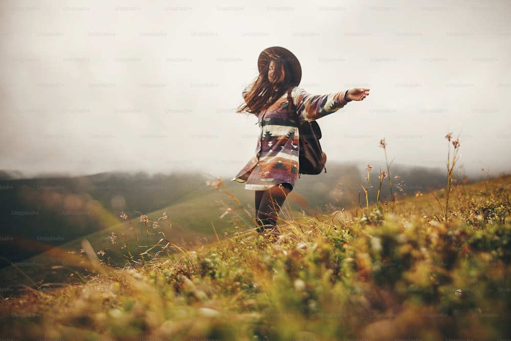 Chica hipster viajera feliz con sombrero, caminando con mochila y sonriendo en montañas ventosas en nubes. espacio para el texto. Increíble momento emocional atmosférico. Concepto de viaje y pasión por los viajes