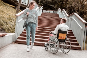 Unusual problem. Serious young woman passing the invalid man sitting in the wheelchair in front of the stairs