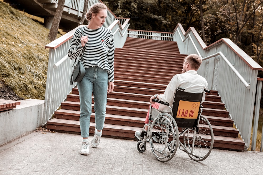 Problema inusual. Mujer joven seria que pasa al hombre inválido sentado en la silla de ruedas frente a las escaleras