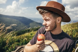 stylish traveler man in hat eating apple on top of sunny mountains. travel and wanderlust concept. space for text. happy hipster traveling and camping. summer vacation