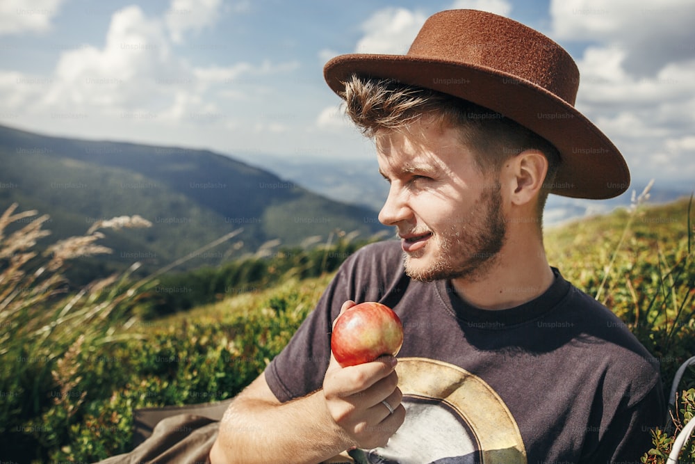 stylish traveler man in hat eating apple on top of sunny mountains. travel and wanderlust concept. space for text. happy hipster traveling and camping. summer vacation