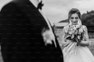 Gorgeous bride with modern bouquet looking at stylish groom and smiling outdoors. Sensual wedding couple posing. Romantic moments of newlyweds. Wedding photo
