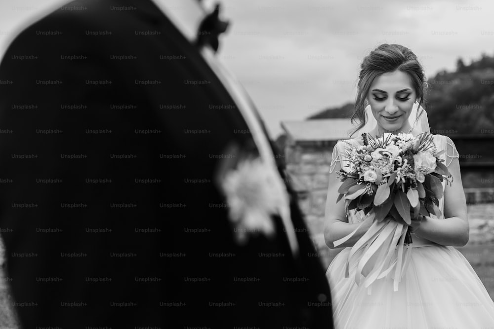 Wunderschöne Braut mit modernem Blumenstrauß, die stilvollen Bräutigam betrachtet und im Freien lächelt. Sinnliches Hochzeitspaar posiert. Romantische Momente des Brautpaares. Hochzeitsfoto