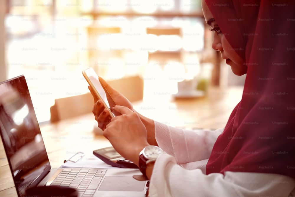 Attractive young Asian wearing red hijab  using her mobile smartphone at workspace desk.