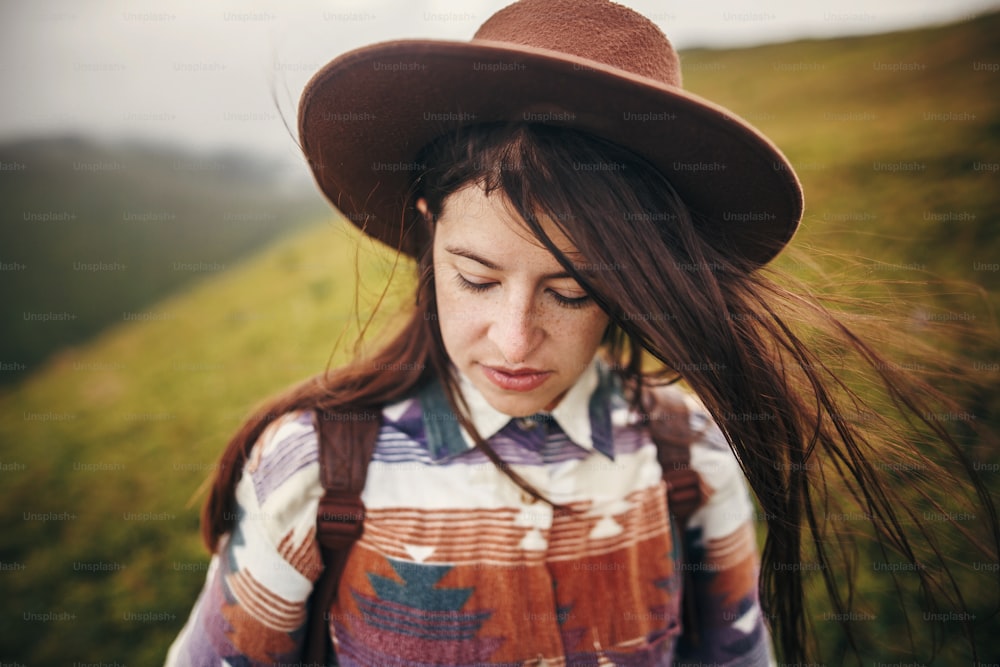 happy traveler hipster girl in hat with windy hair, relaxing in sunny mountains. space for text. atmospheric emotional moment. travel and wanderlust concept.  woman portrait