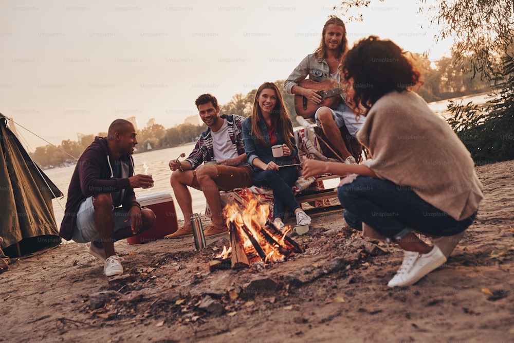 Group of young people in casual wear smiling while enjoying beach party near the campfire