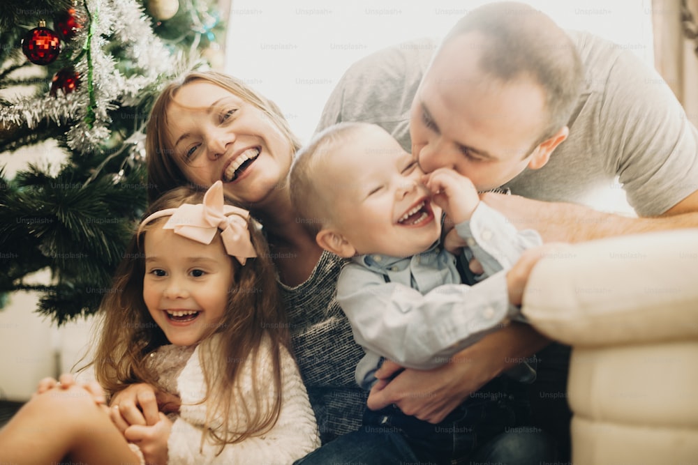 Cheerful adult parents with little boy and girl having fun and embracing on floor in living room with ChristmasÂ tree