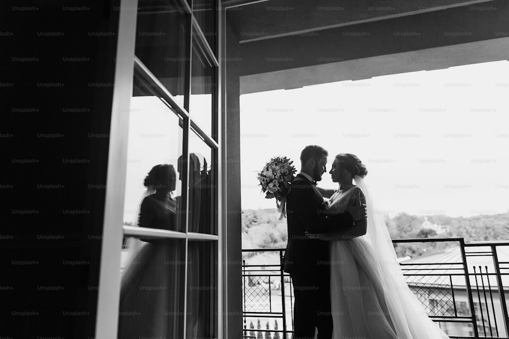 Gorgeous bride and stylish groom silhouettes at window light. Sensual wedding couple embracing. Romantic moments of newlyweds. Creative wedding photo. Copy space