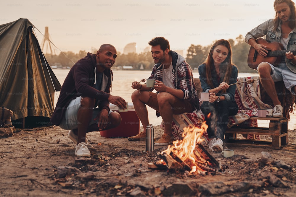 Gruppo di giovani in abbigliamento casual sorridente mentre si gode la festa in spiaggia vicino al fuoco