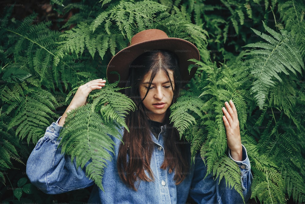 stylish hipster girl in hat sitting in fern bushes, among fern leaves in forest. creative portrait of woman traveler. environmental concept. space for text. atmospheric moment