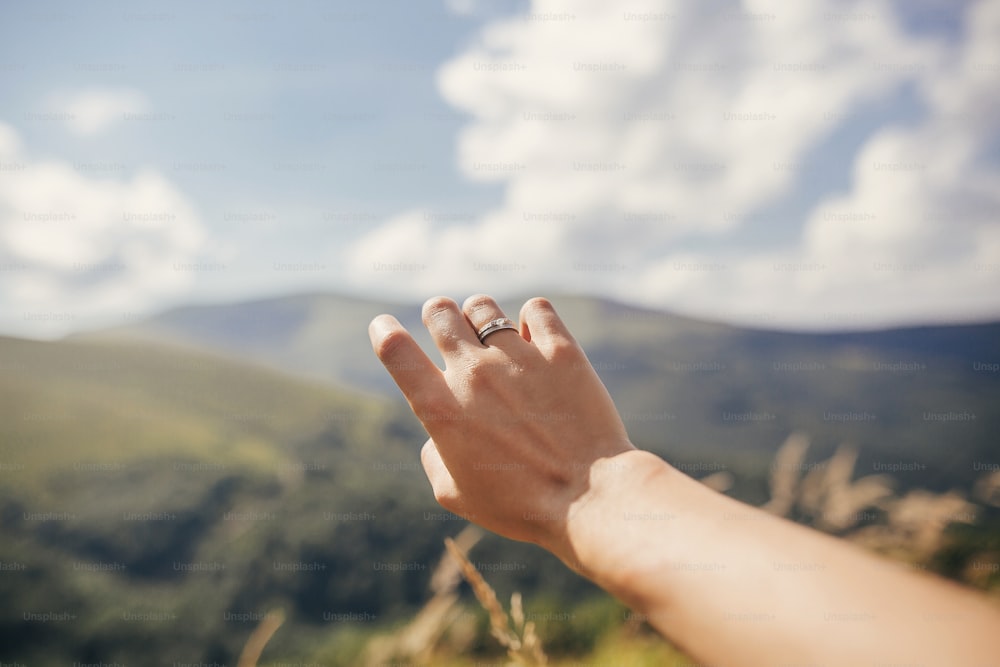 traveler hand reaching out to mountains with engagement ring. girl hand on background of sunny mountains and sky. travel and wanderlust concept. summer vacation