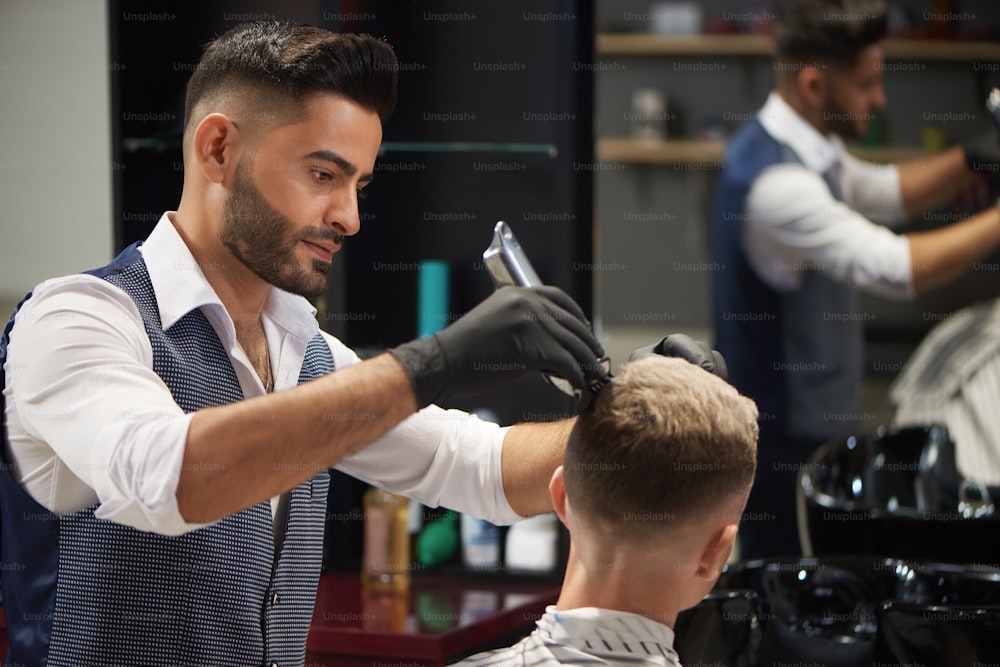 Concentrated male hairdresser holding trimmer in hands and trimming haircut of client. Bearded master wearing white shirt, vest and black gloves, while working in barbershop.