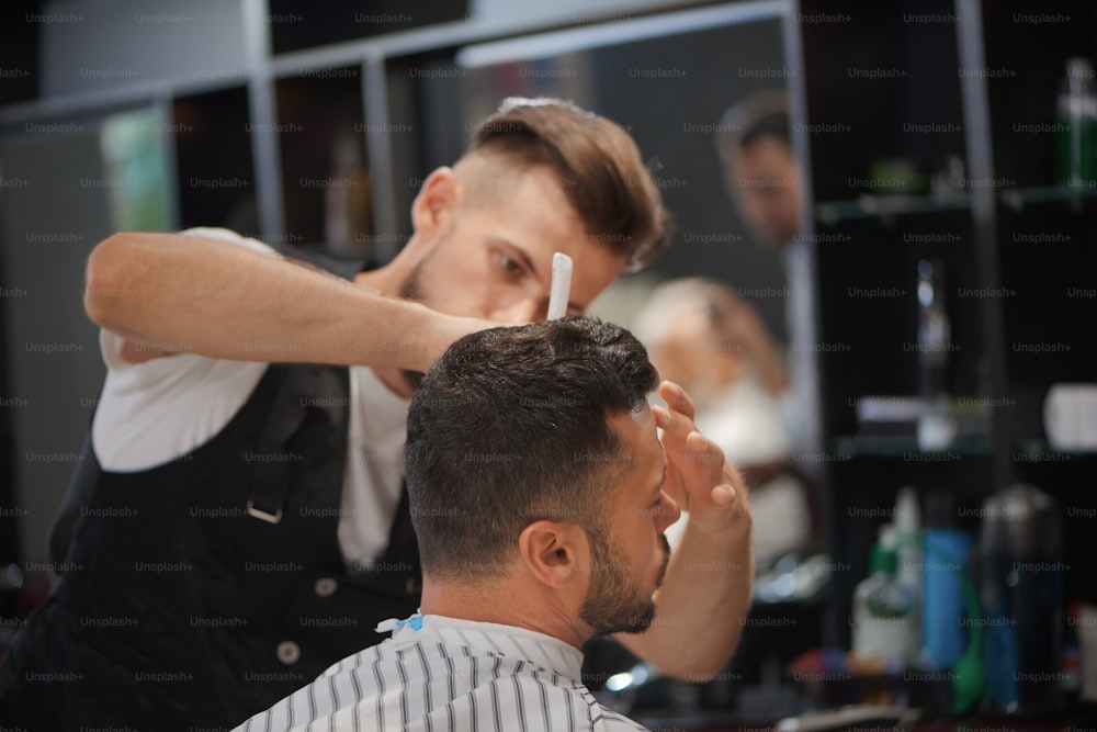 Confident male hairdresser styling man's beard and stylish haircut. Bearded client waiting while barber grooming his hair. Concept of barbershop, beauty and hair care.