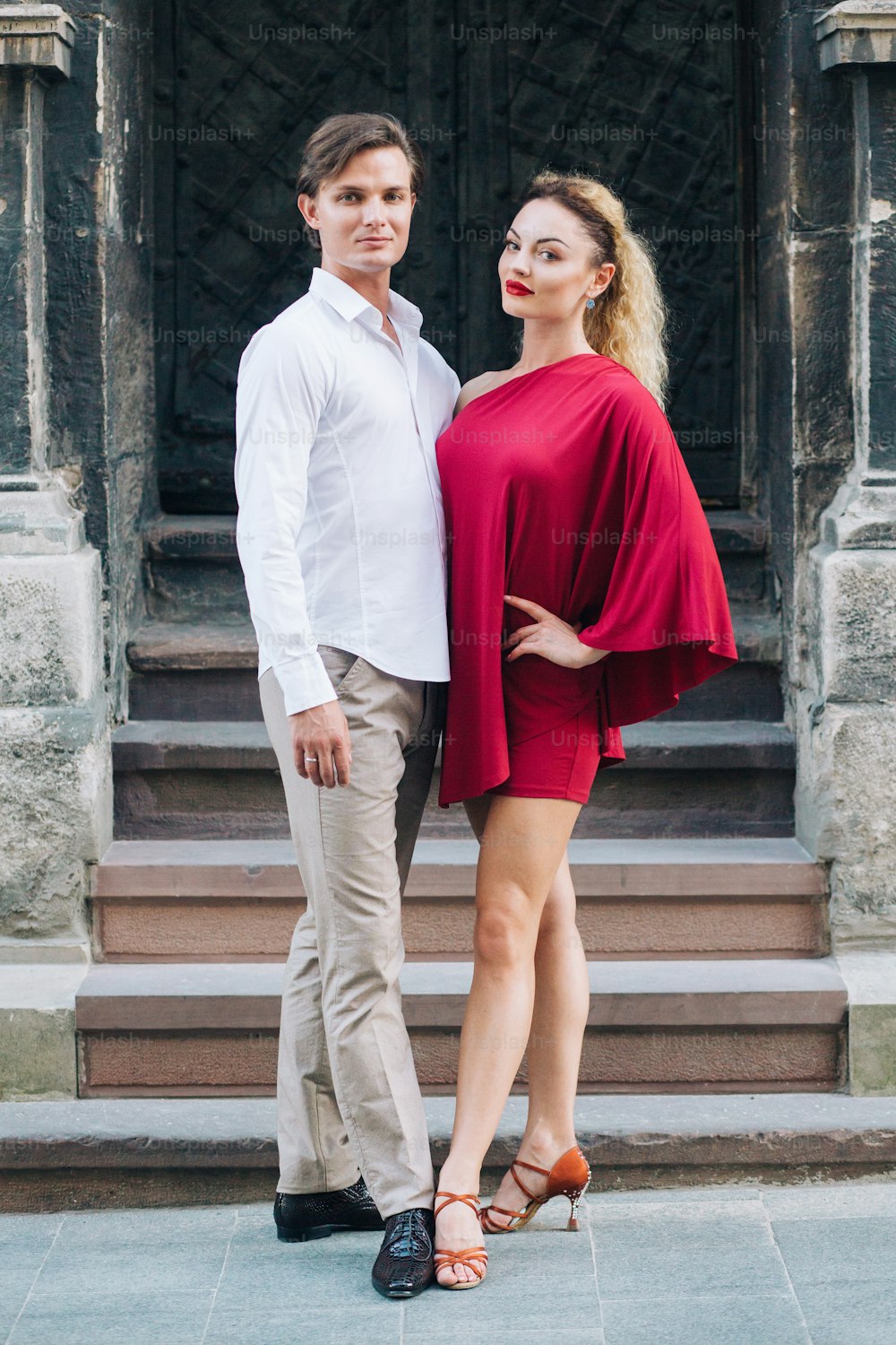 beautiful happy couple in love posing at old building in sunny street. stylish hipster groom and blonde bride in red dress gently hugging, professional dancers. romantic moments in summer