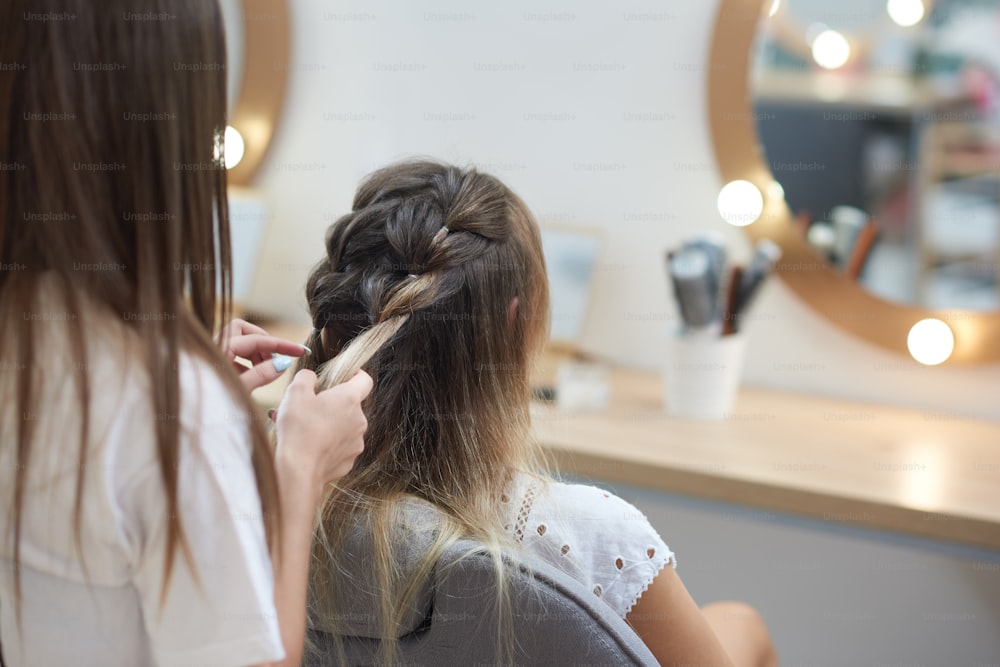 View from back of female hairdresser braiding two spikelets to young long haired client in beauty salon. Professional hair cutter in process of doing trendy hairstyle. Concept of beauty and fashion.