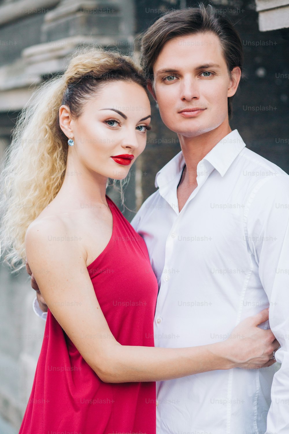 日当たりの良い通りの古い建物でポーズをとる愛の美しい幸せなカップル。スタイリッシュなヒップスターの花婿と赤いドレスを着たブロンドの花嫁が優しく抱きしめる、プロのダンサー。夏のロマンチックなひととき