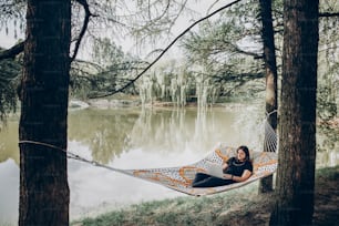 Freelance hipster élégant avec ordinateur portable se relaxant dans un hamac dans un parc d’été ensoleillé. fille regardant l’ordinateur portable, travaillant et souriant. Concept de freelance, travaillant à l’extérieur. vacances d’été, espace pour le texte