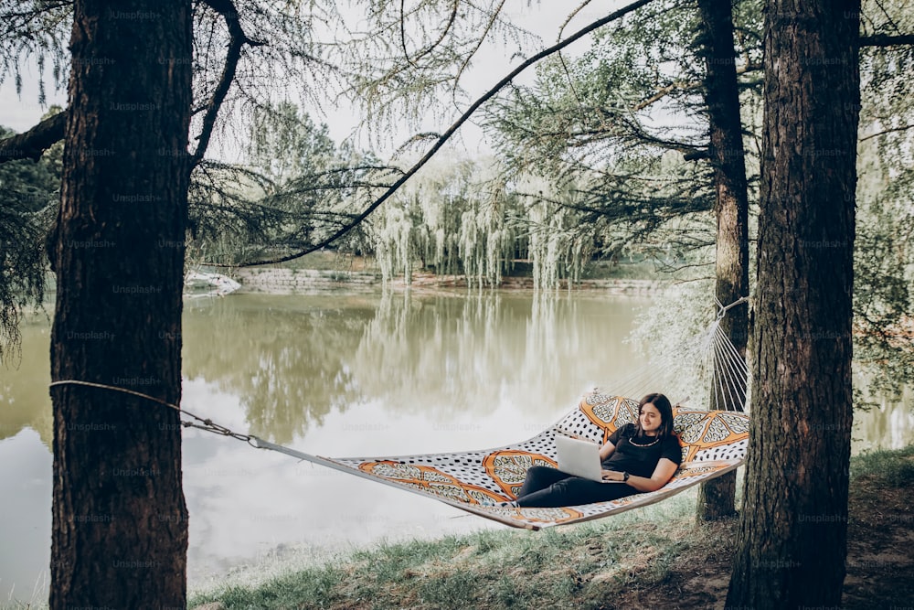 stylish hipster freelancer with laptop relaxing in hammock in sunny summer park. girl looking at laptop, working and smiling. freelance concept, working outdoors. summer vacation, space for text