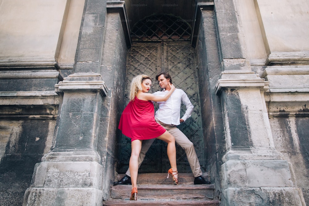 Schönes, glückliches verliebtes Paar, das sich im alten Gebäude in der sonnigen Straße umarmt und tanzt. Stilvoller Hipster-Bräutigam und blonde Braut im roten Kleid tanzen, professionelle Tänzer. Romantische Momente