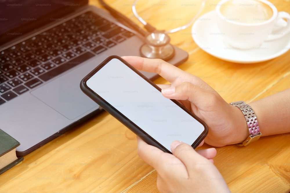 Manos femeninas sosteniendo un teléfono inteligente móvil con pantalla en blanco en el escritorio de la oficina.