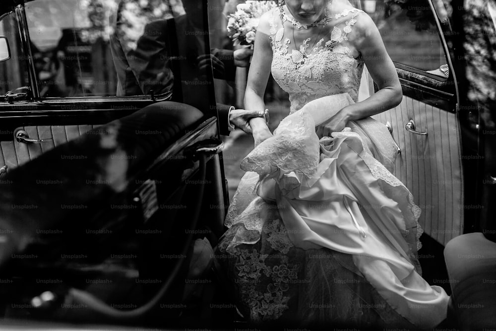 Stylish bride in vintage white wedding dress getting into black limousine with groom, happy couple sitting in car before wedding ceremony