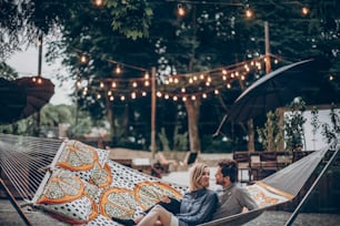stylish hipster couple cuddling and relaxing in hammock under retro lights in evening summer park. man hugging woman and resting in forest. space for text. rustic love concept