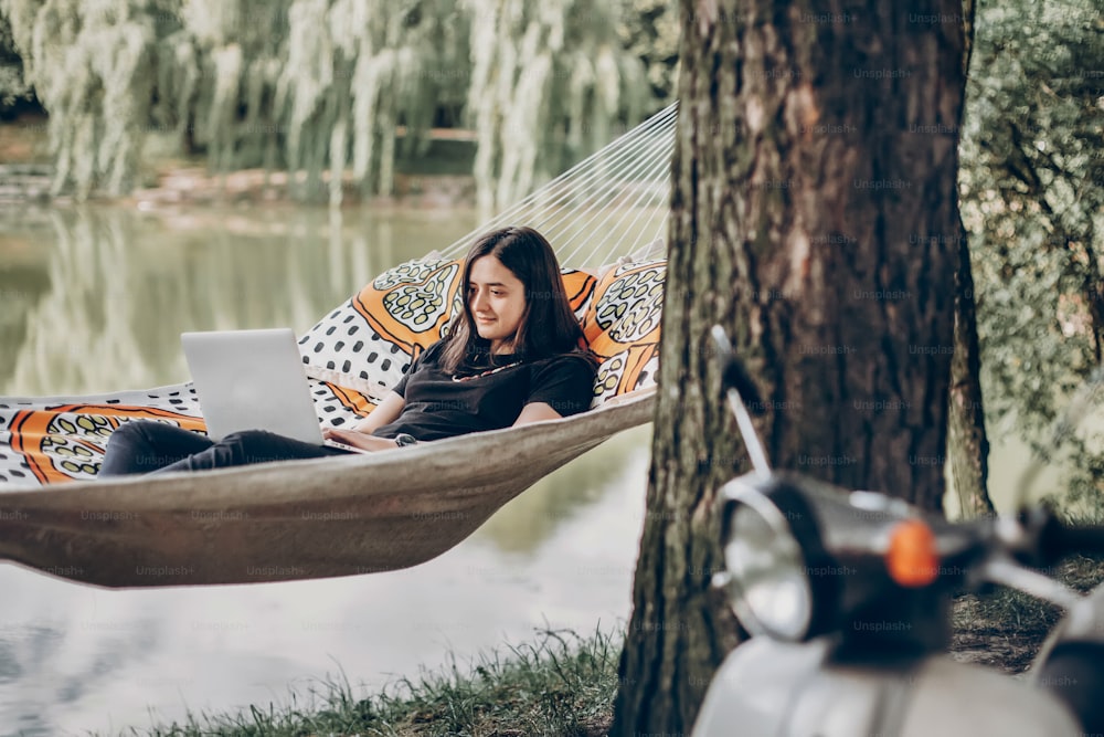 Joven trabajadora independiente que trabaja en una computadora portátil mientras está acostada en una hamaca cerca de un lago, mujer hipster descansando en la naturaleza cerca de una motocicleta italiana retro