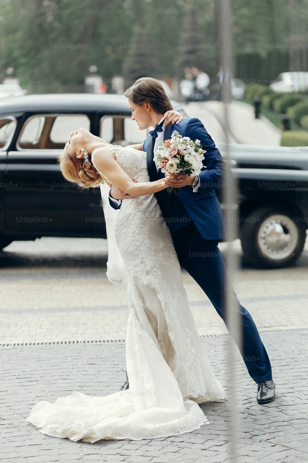Romantic bride and groom portrait after wedding ceremony, happy newlywed couple dancing outdoors, luxury car in background, happy groom hugging bride