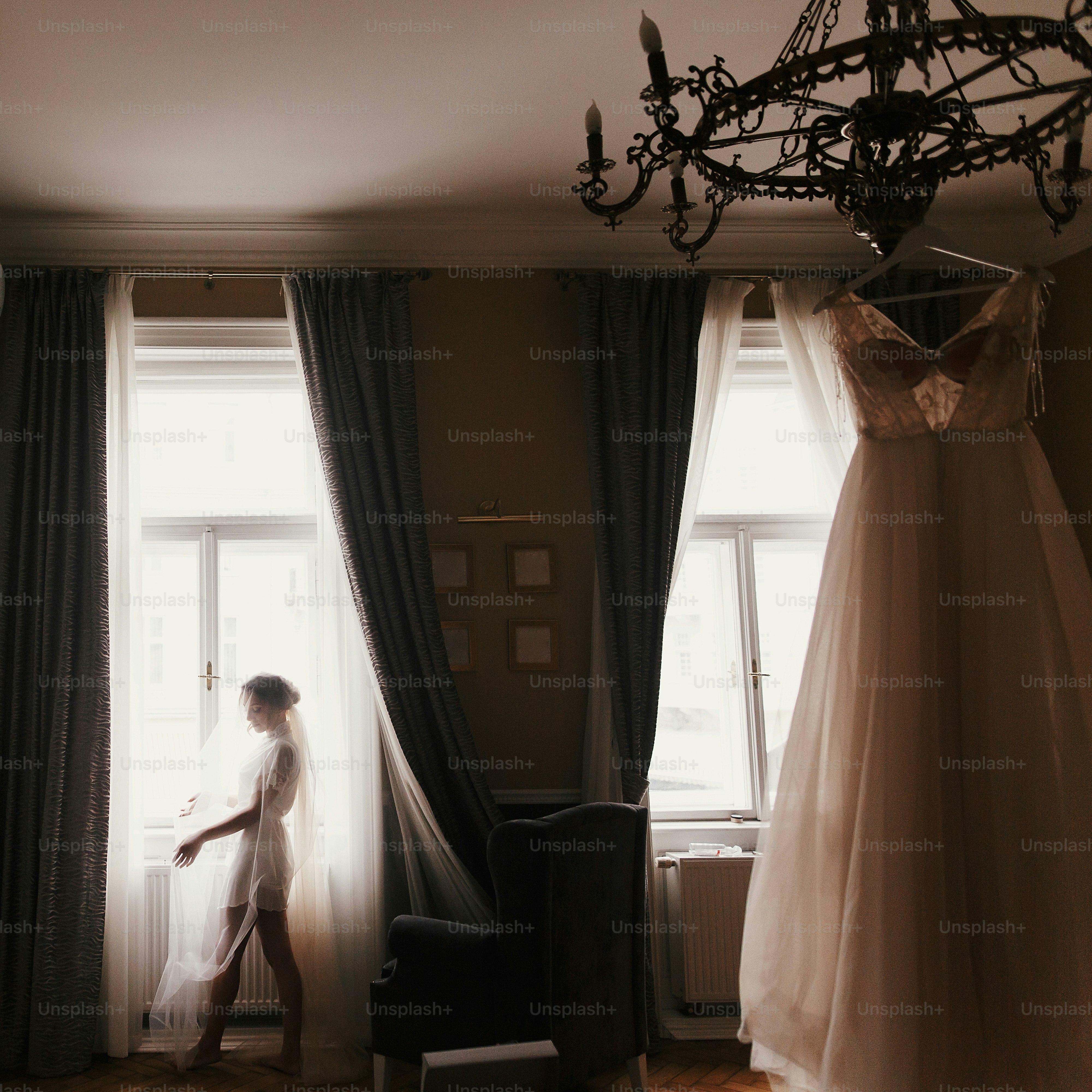 Splendida silhouette della sposa in piedi alla luce della finestra, guardando l'elegante abito da sposa, appeso al lampadario, al mattino. Sposa e il suo bellissimo abito da sposa. Donna che si prepara