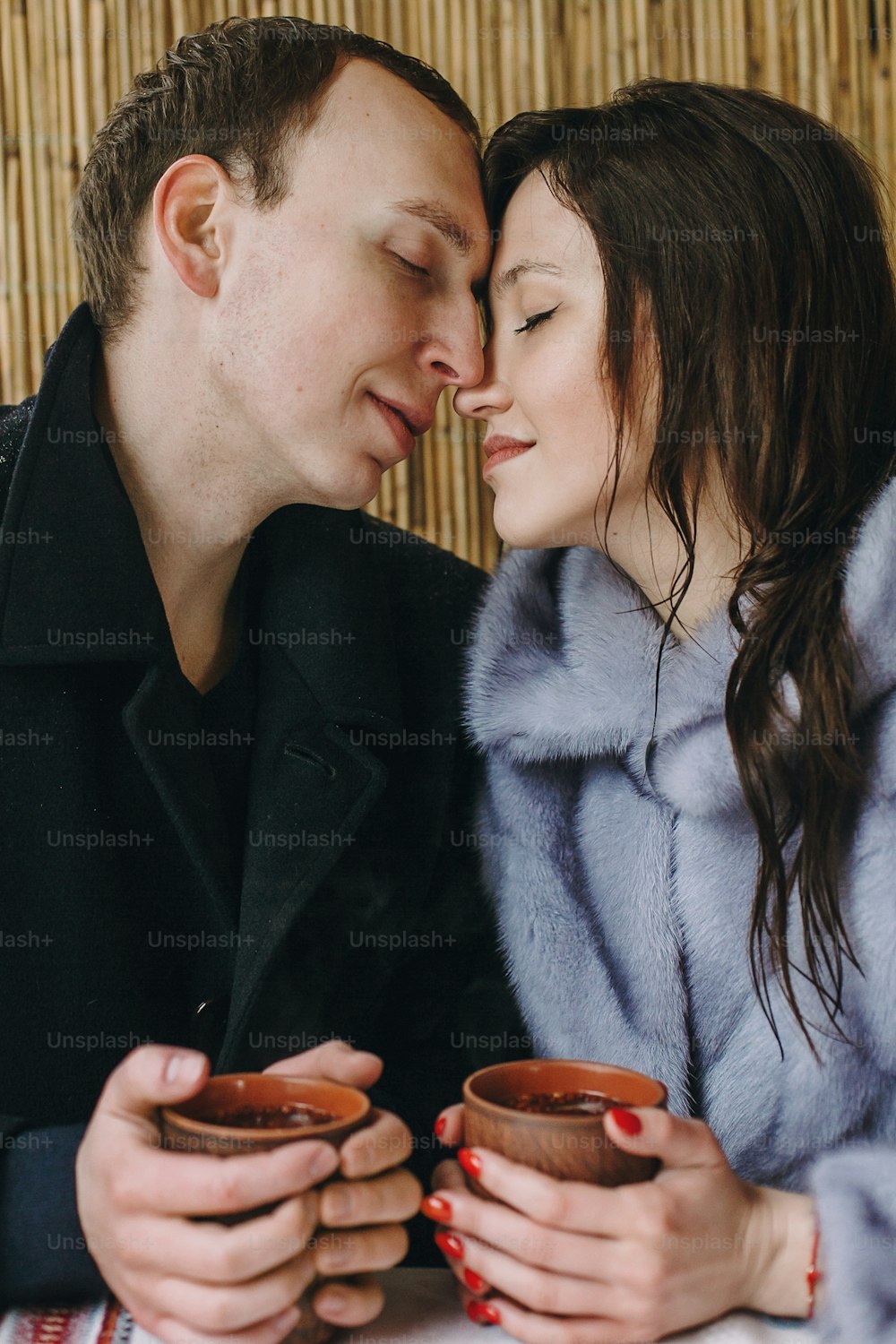 Couple élégant tenant du thé chaud dans des tasses et s’embrassant sur un porche en bois dans les montagnes enneigées d’hiver. Famille romantique heureuse avec des boissons étreintes. Escapade de vacances ensemble