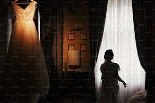 Splendida silhouette della sposa in piedi al vestito da sposa elegante, appesa al lampadario, al mattino alla luce della finestra. Sposa e il suo bellissimo abito da sposa. Donna che si prepara