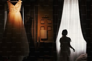 Gorgeous bride silhouette standing at stylish wedding dress, hanging on chandelier, in the morning at window light. Bride and her beautiful wedding gown. Woman getting ready