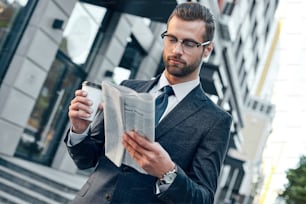 Jeune homme d’affaires en costume et lunettes tenant un gobelet en papier et lisant un journal d’affaires dans ses mains