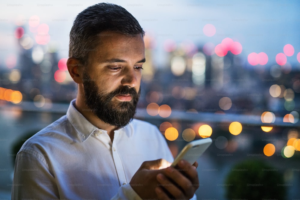 A portrait of businessman with smartphone standing against night London rooftop view panorama at sunset, text messaging. Copy space.