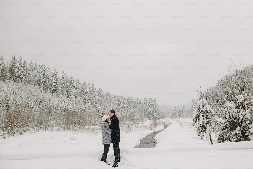 Stylish couple in love holding hands and embracing in snowy mountains. Happy family gently hugging and kissing in winter mountains and forest. Space for text
