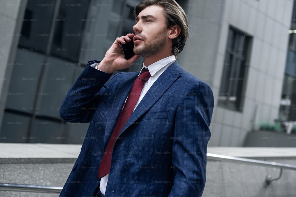 Confident businessman. Confident young man in full suit is talking by his smart phone and looking away while standing outdoors with cityscape in the background