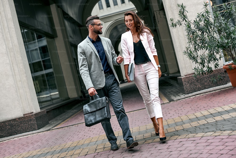 Discussing project. Two young business people walking outside on the city street talking smiling happy man carrying briefcase