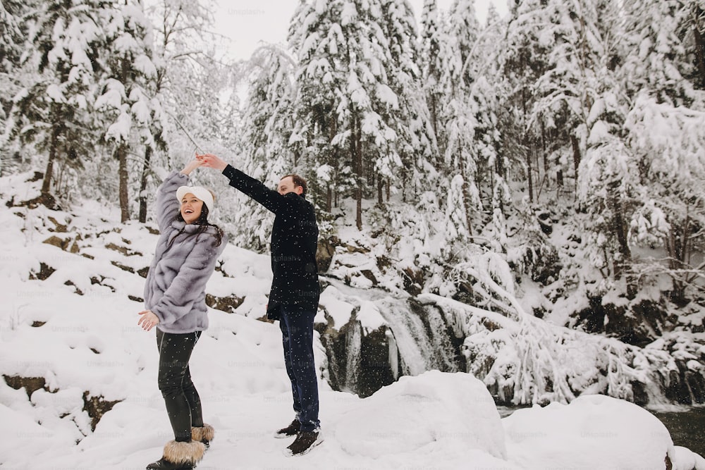 Stylish couple having fun in winter snowy mountains. Happy romantic man and woman in luxury clothes smiling and playing at waterfall in snow. Holiday getaway together.