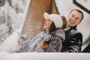 Couple élégant jouant avec la neige dans une cabane en bois sur fond de montagnes enneigées d’hiver. Famille heureuse s’amusant et souriant dans la neige au chalet. Escapade de vacances ensemble