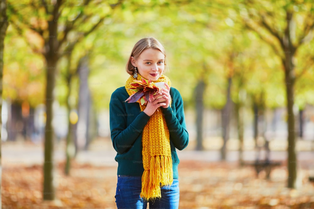 Muchacha joven feliz en bufanda amarilla con café para ir a caminar en el parque de otoño en un día brillante de otoño