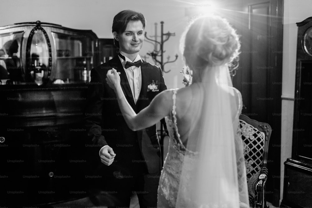 幸せな新婚カップル、ロマンチックな笑顔の花婿は、ホテルで屋内でエレガントな白いウェディングドレスを着たゴージャスな花嫁を見ています
