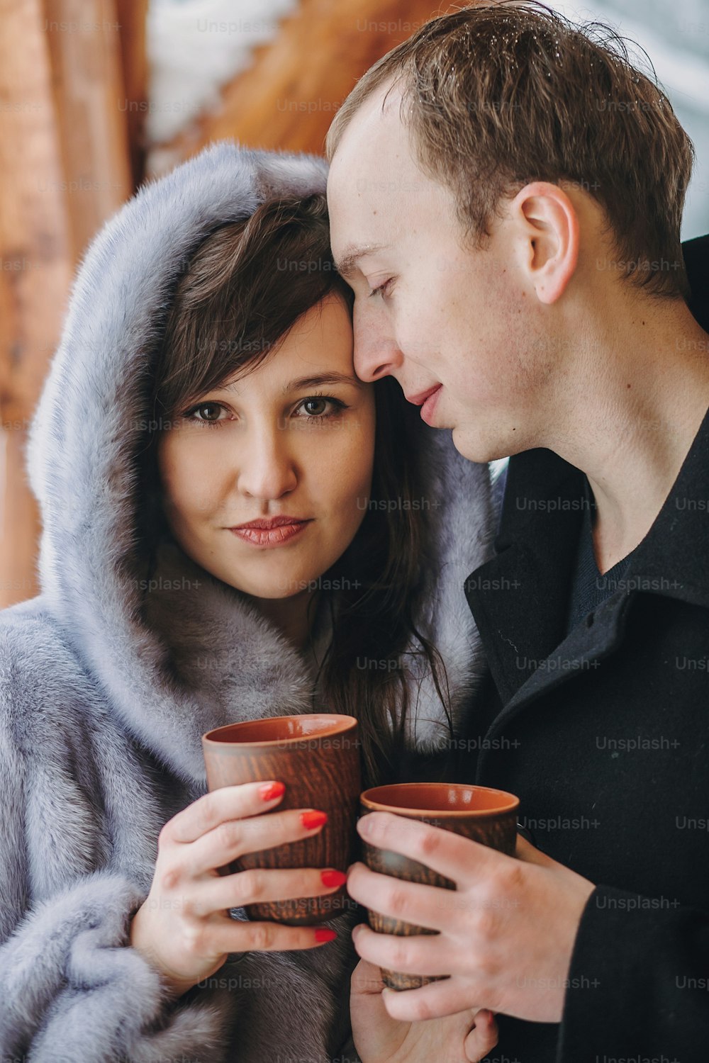 Stylish couple holding hot tea in cups and embracing on wooden porch in winter snowy mountains. Happy romantic family with drinks hugging. Holiday getaway together.