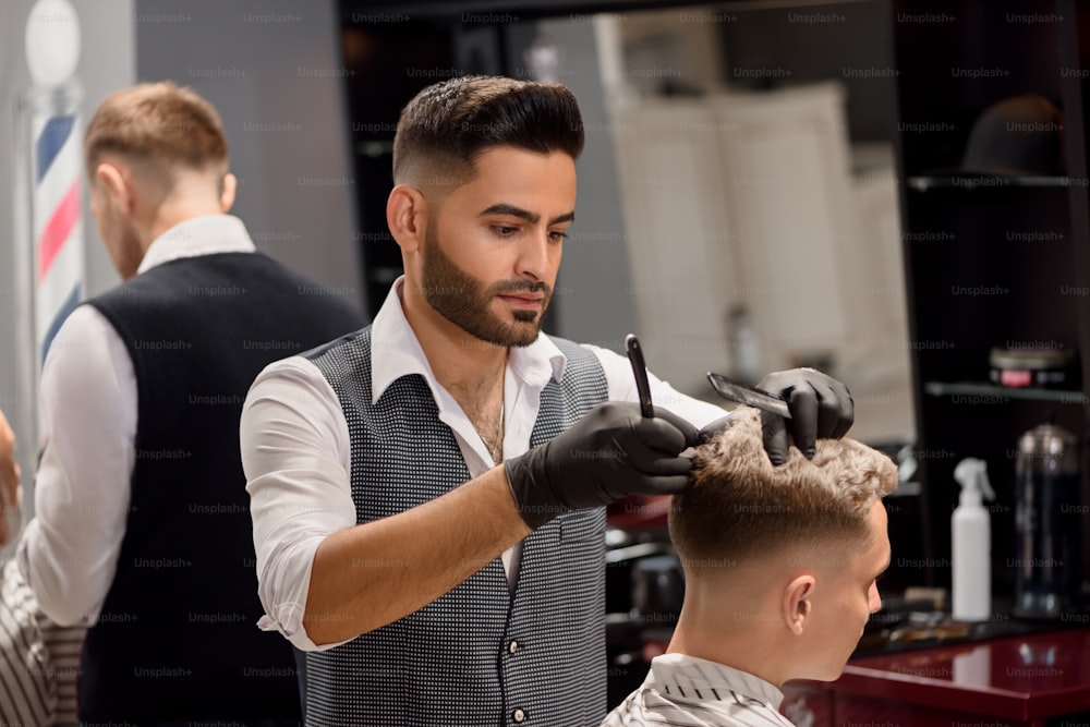 Concentrated hairdresser making professional and stylish haircut to client. Serious master using black comb and razor. Bearded barber wearing white classic shirt and grey vest.