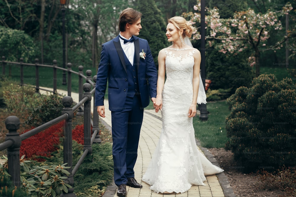 stylish bride and groom relaxing in park.  happy luxury wedding couple walking and smiling among trees. man in blue suit and woman in white dress at reception outdoors